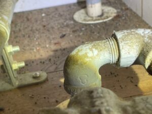 茨城県つくば市の古いアパートにて、キッチン下の収納内部を調査。ガスの元栓が通る床の隙間周辺に、大量のゴキブリの糞と卵鞘が散乱している様子を捉えた現場写真